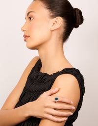 Featuring a statement ring with plated material and cubic zirconia border surrounding an oval cut center stone in deep blue on finger view - link has visual effect only
