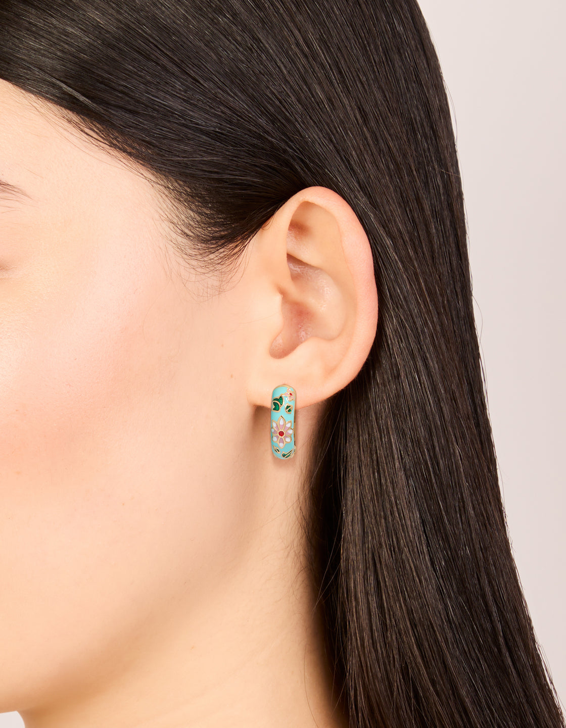 Crafted from enamel, these thick huggie earrings feature floral and leaf print in turquoise, gold, pink, and green with huggie closure on ear view
